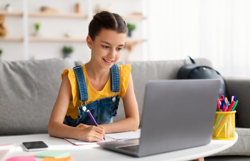 Cheerful teen sitting at desk, using laptop, drawing in copybook