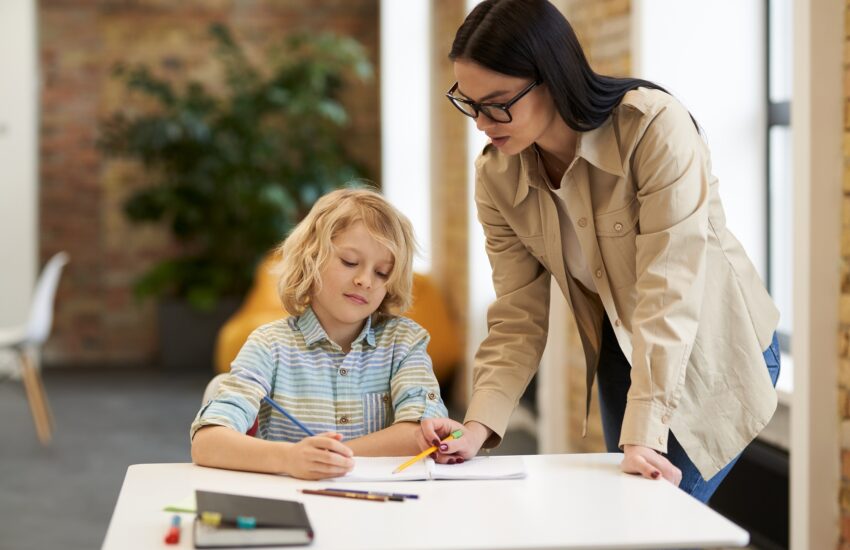 Supportive young female teacher in glasses helping little boy with the task. Kid studying in